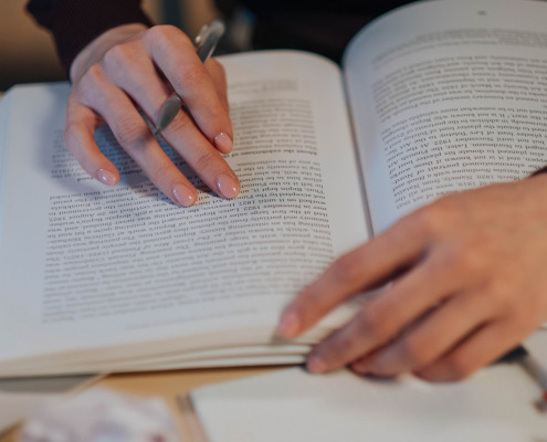 Zwei Hände liegen auf einem offenen Buch auf, in der rechten Hand befindet sich zwischen den Fingern ein Stift.